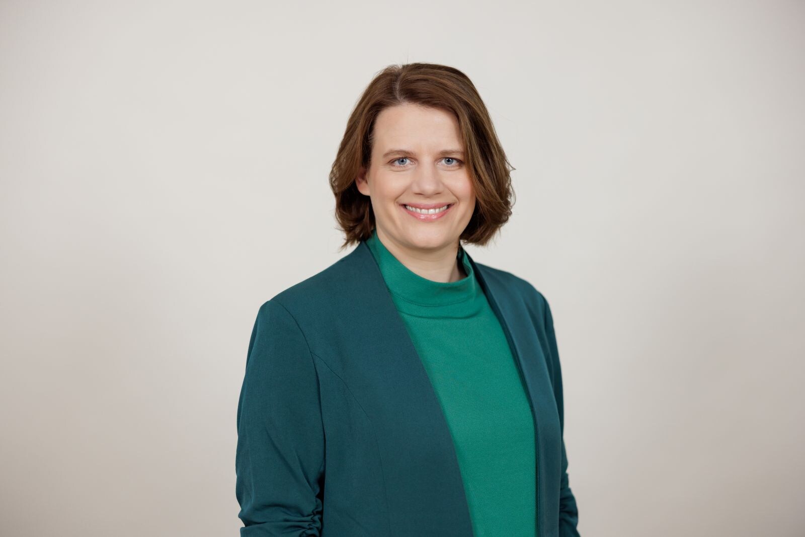 Eine Frau mit kurzen braunen Haaren trägt ein grünes Oberteil und einen Blazer und lächelt vor neutralem Hintergrund in die Kamera.