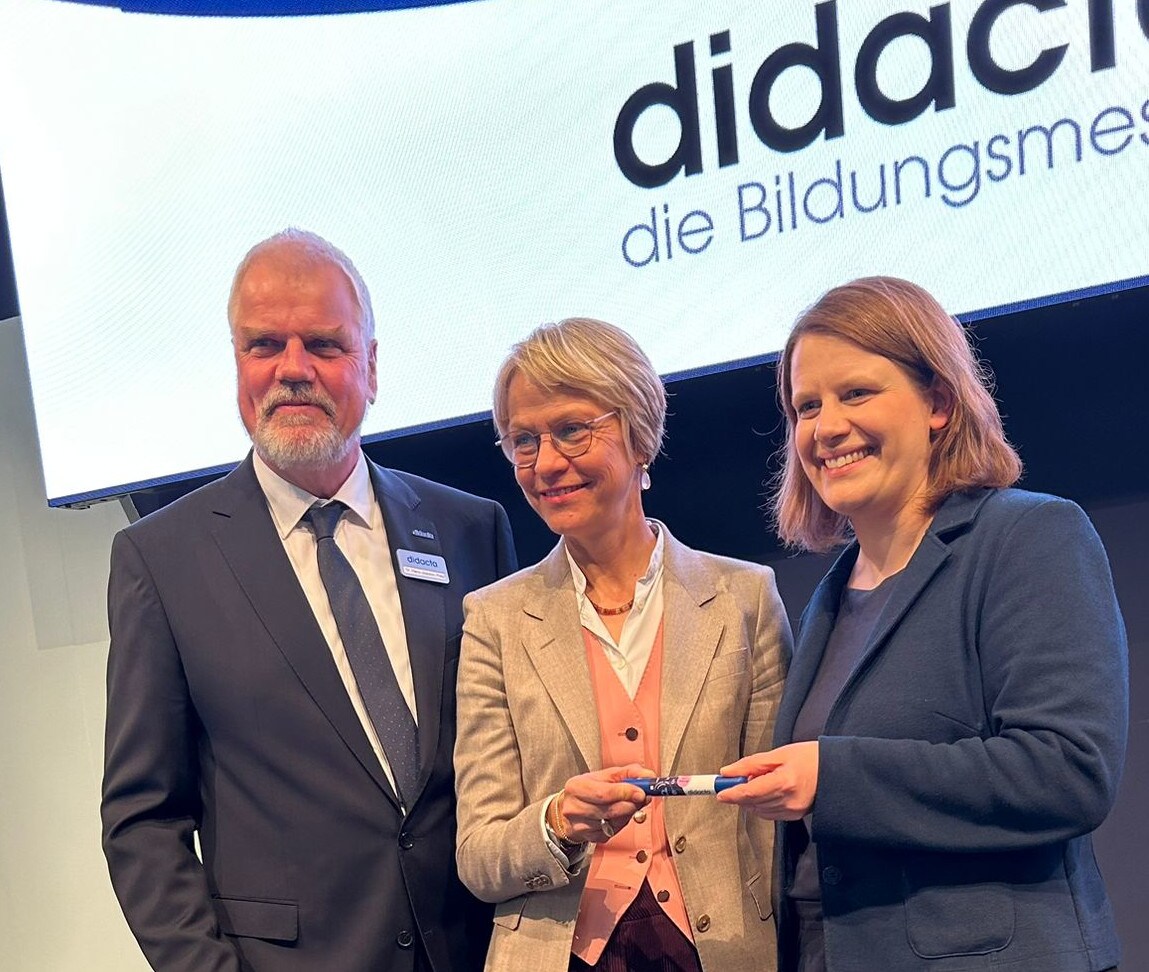 Drei Personen stehen auf einer Bühne vor einer didacta-Leinwand und überreichen gemeinsam symbolisch einen Gegenstand.
